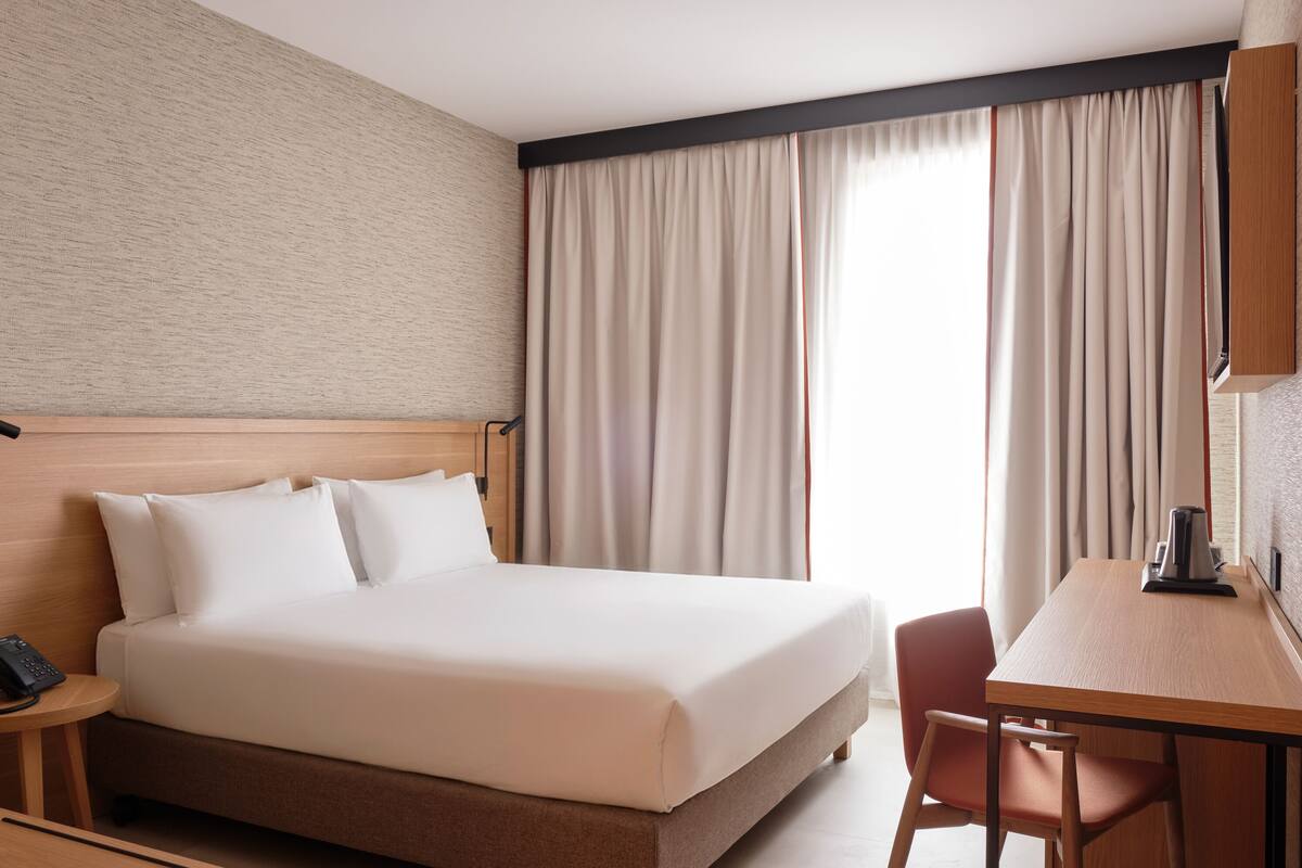 A hotel room with a bed, a desk, a chair, a telephone, and a window with curtains.