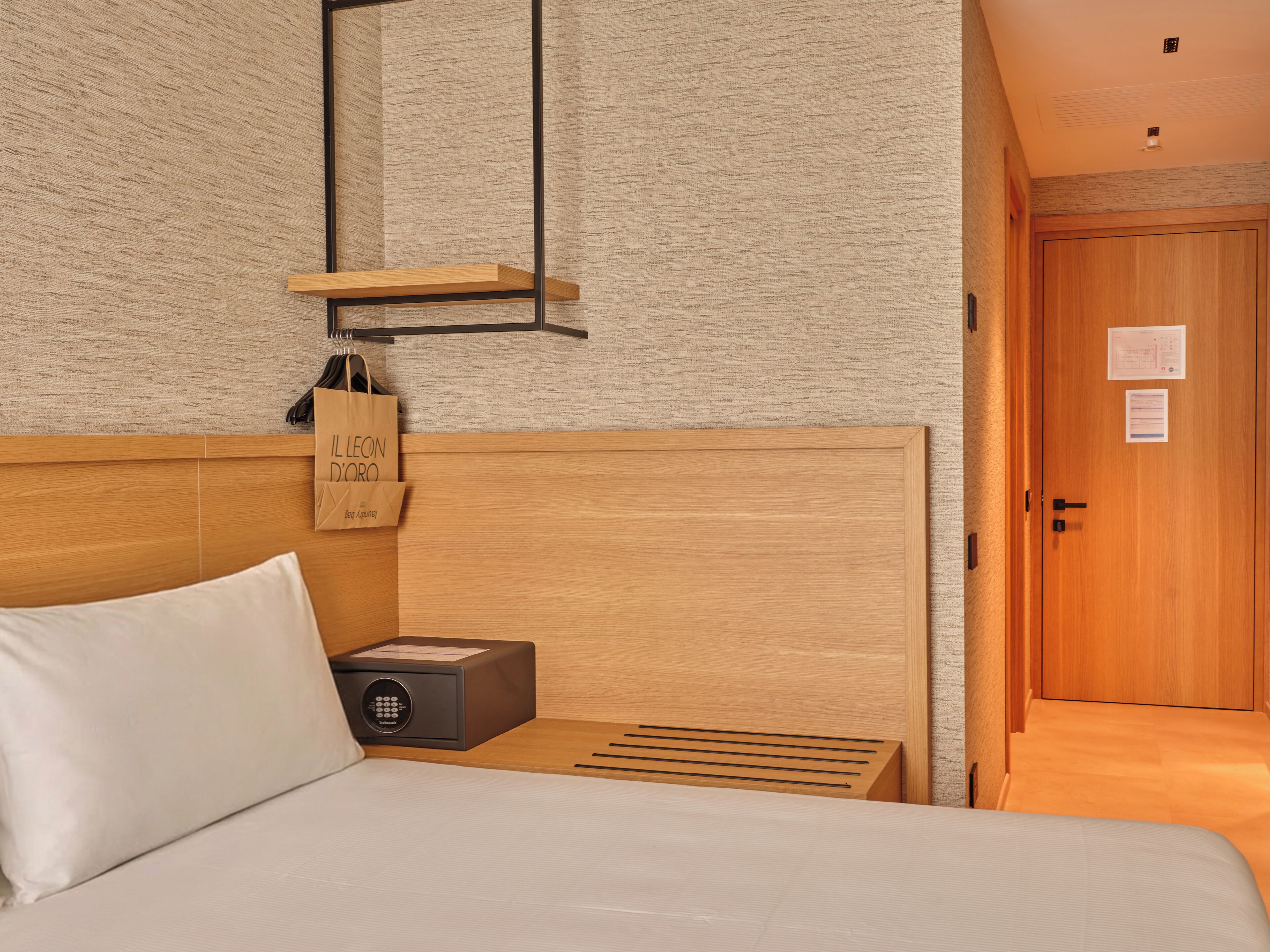 A hotel room with a wooden headboard, a bedside table with a lamp, a nightstand, and a door with a sign.