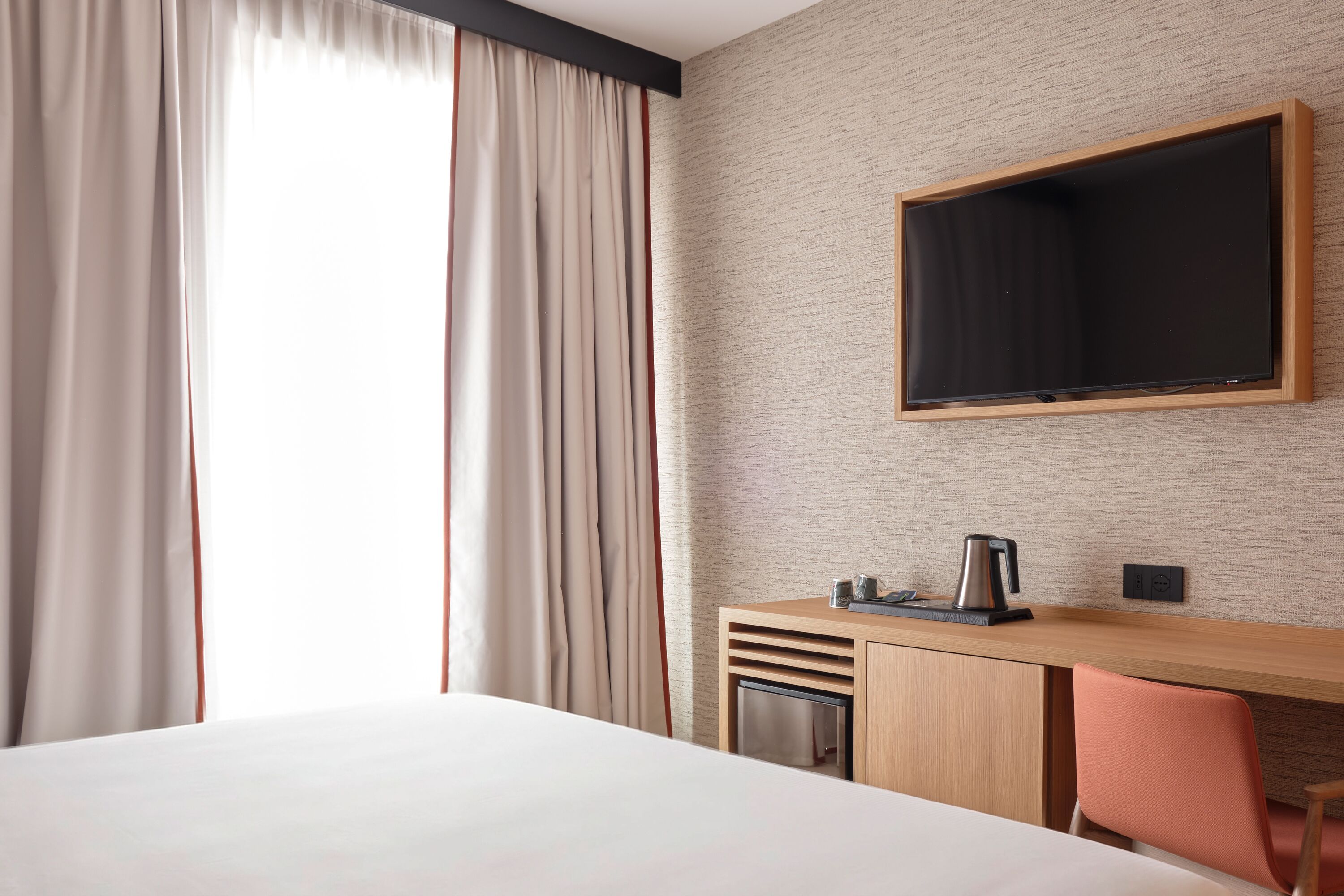 Ein Hotelzimmer mit einem Bett, einem Schreibtisch mit Stuhl, einem an der Wand befestigten Fernseher und einem Fenster mit Vorhängen.