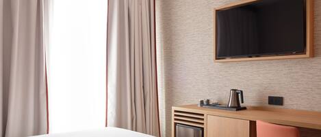 A hotel room with a bed, a desk with a chair, a television mounted on the wall, and a window with curtains.