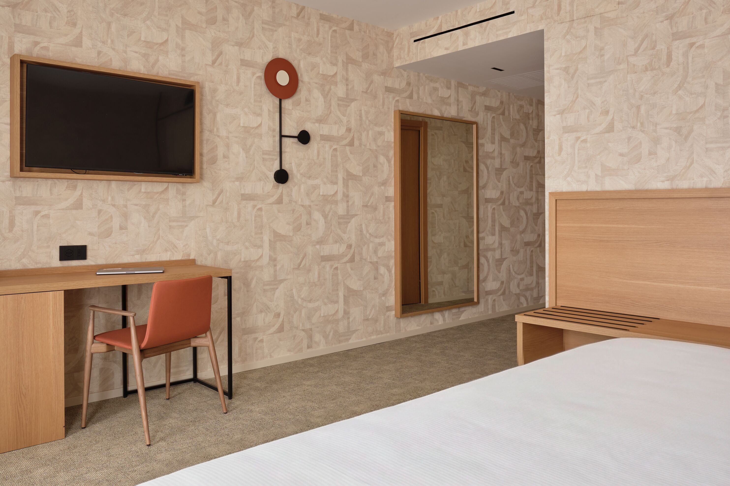A modern hotel room with a wooden desk, a single chair, a large mirror, and a bed.