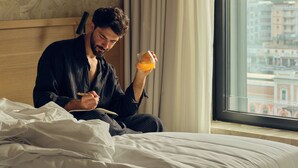 A person sitting on a bed in a hotel room, holding a glass of orange juice, with a view of buildings outside the window. - IL LEON D'ORO HOTEL BARI – BY FARACE HOTELS (Bari)