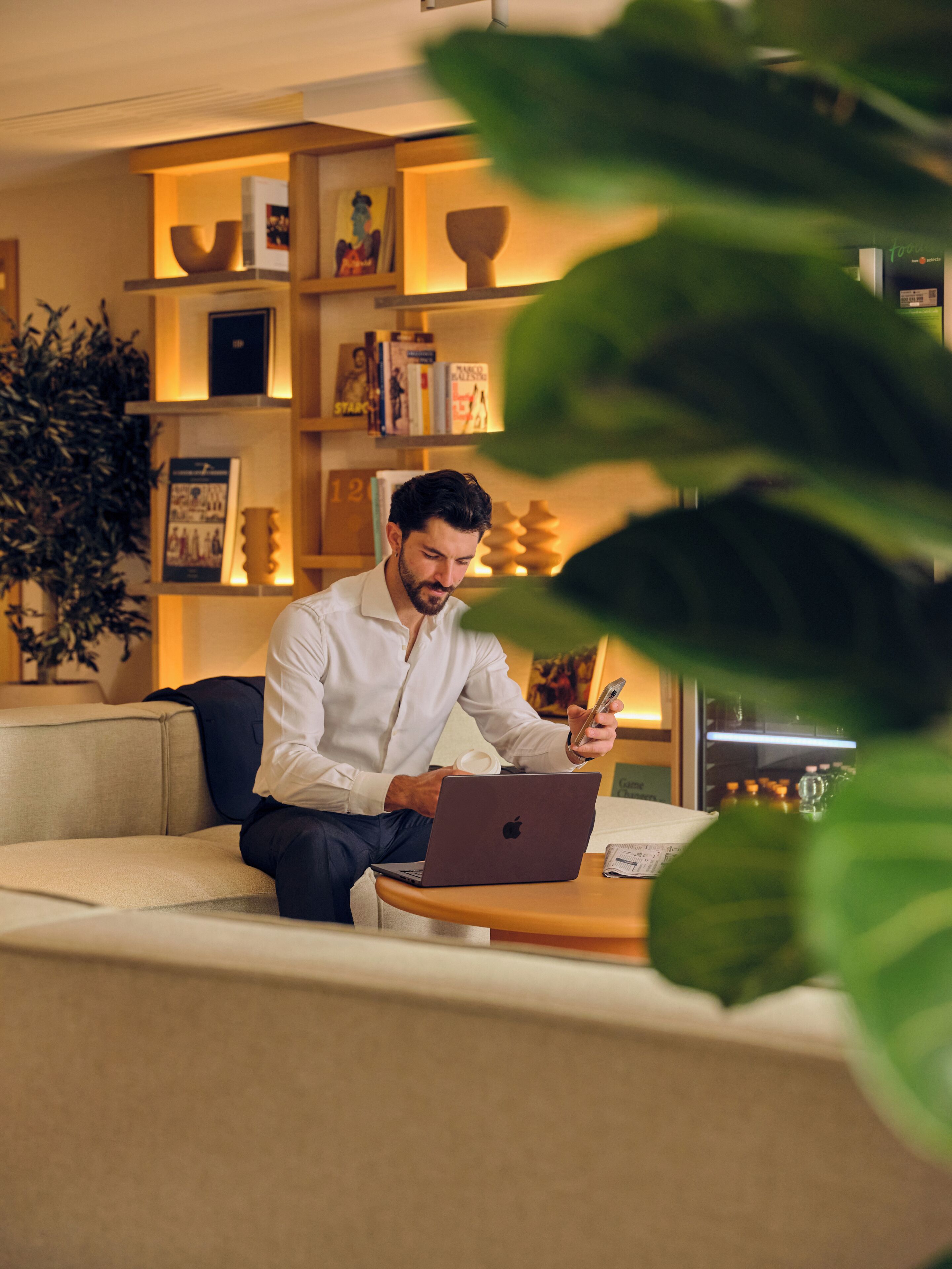 A man sitting on a sofa with a laptop, surrounded by bookshelves and a potted plant.