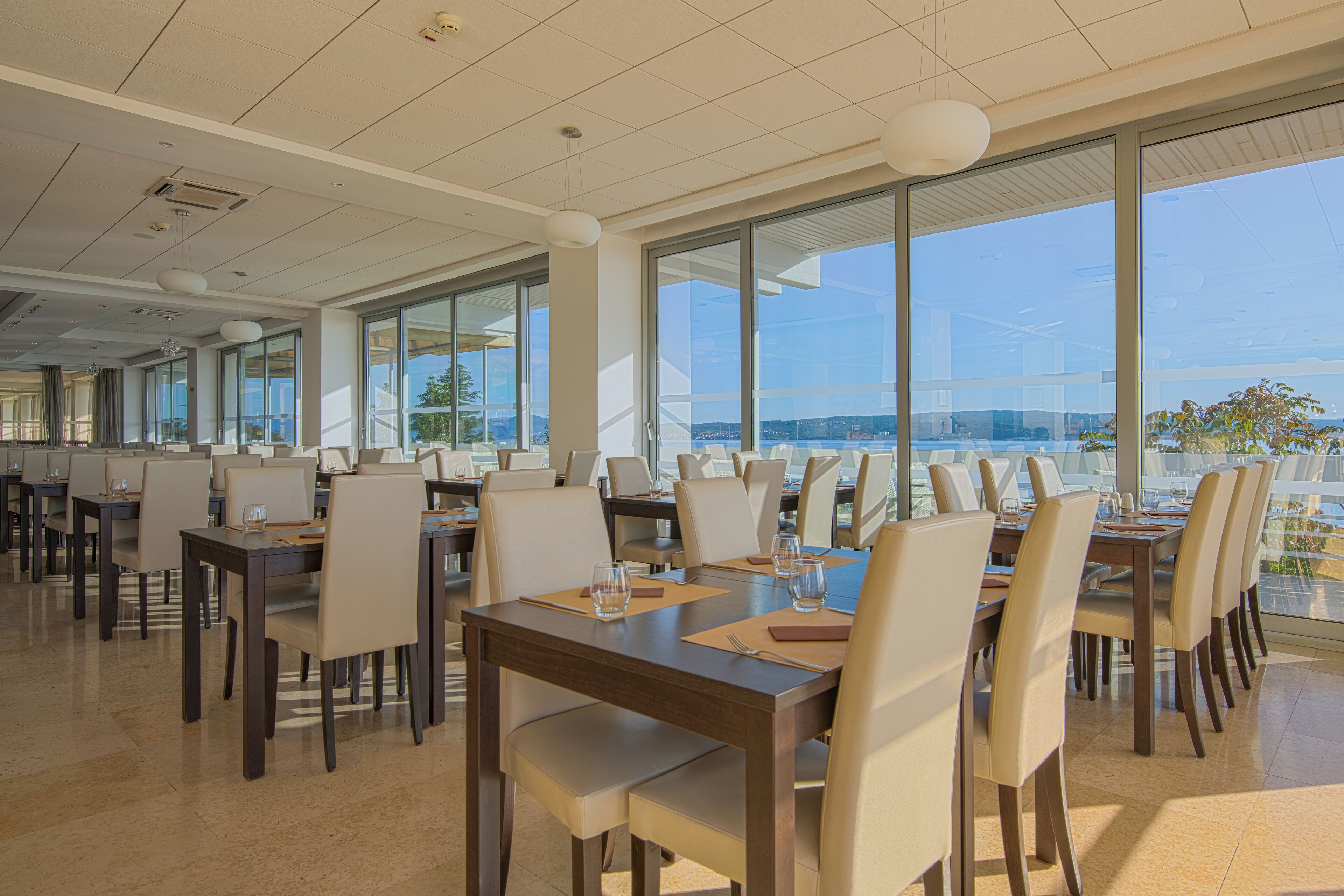 A spacious dining area with multiple tables and chairs, large windows offering a view of the outdoors.