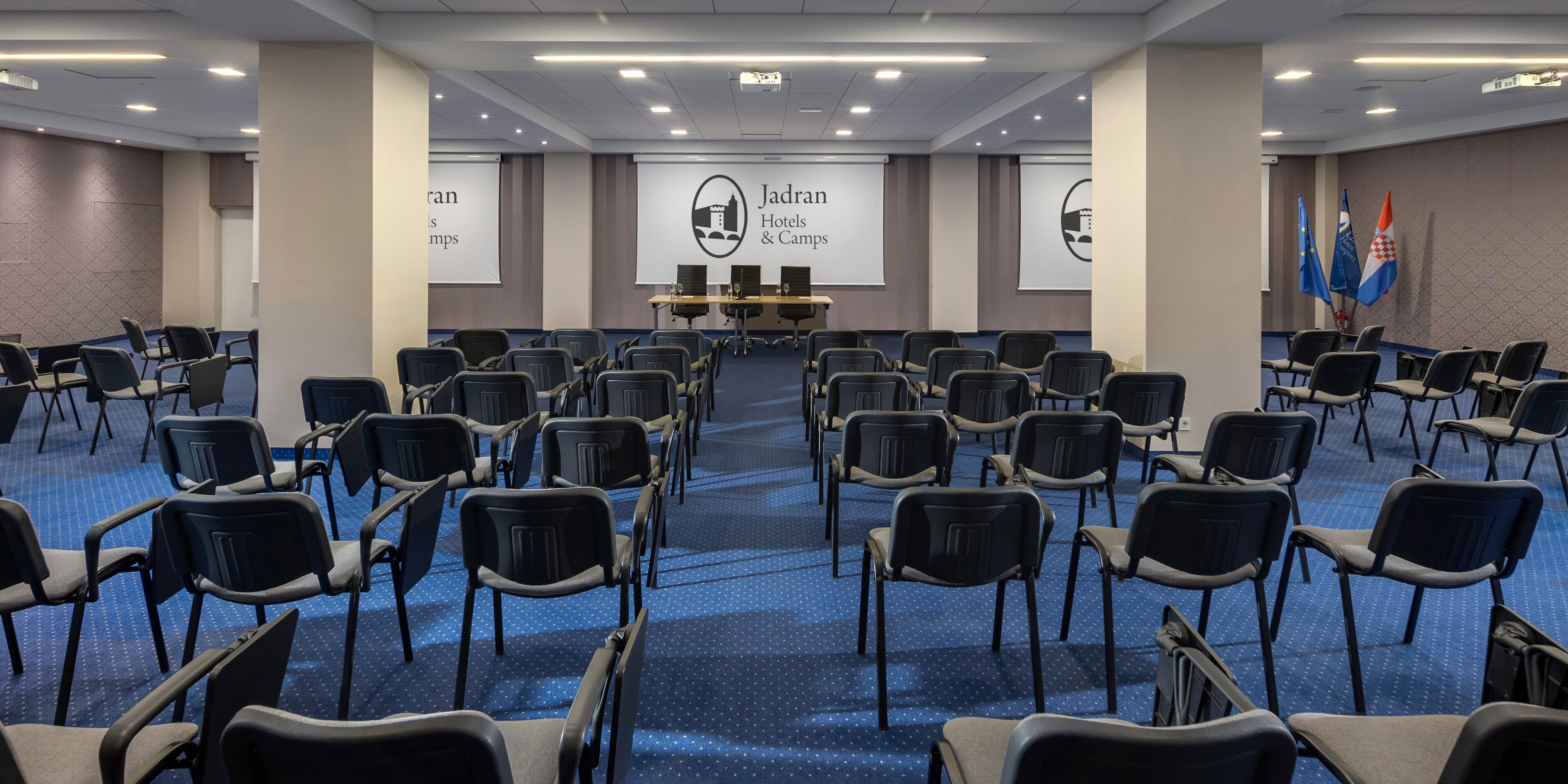Une salle de conférence avec des rangées de chaises noires, une scène avec un pupitre et des banderoles aux couleurs de « Jadran Hotels & Camps ».