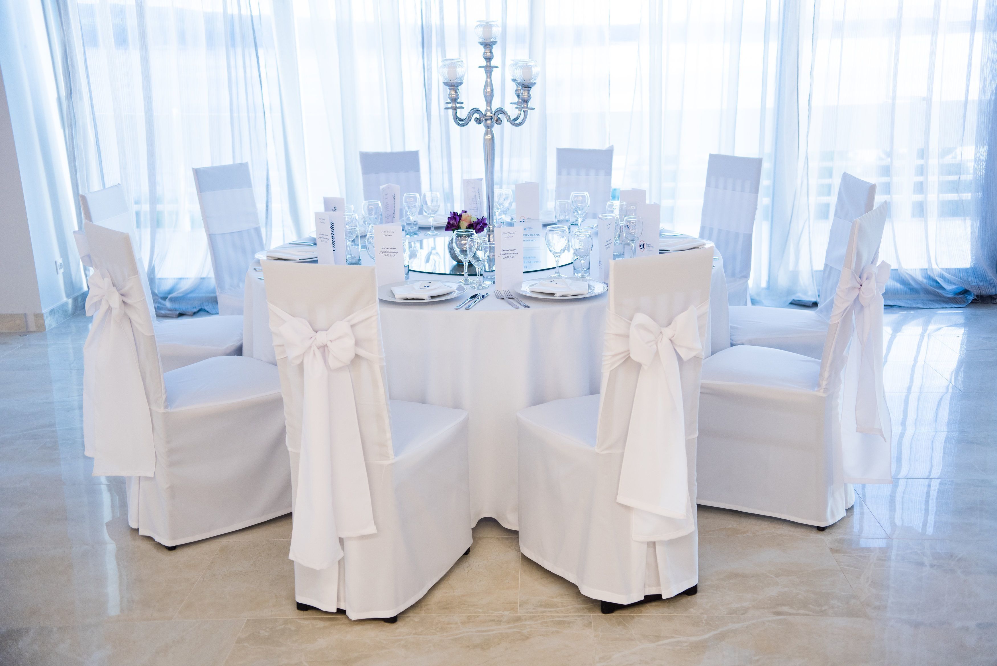 Et rundt bord dækket til en formel lejlighed, med hvide stole og en lysestage som midtpunkt.