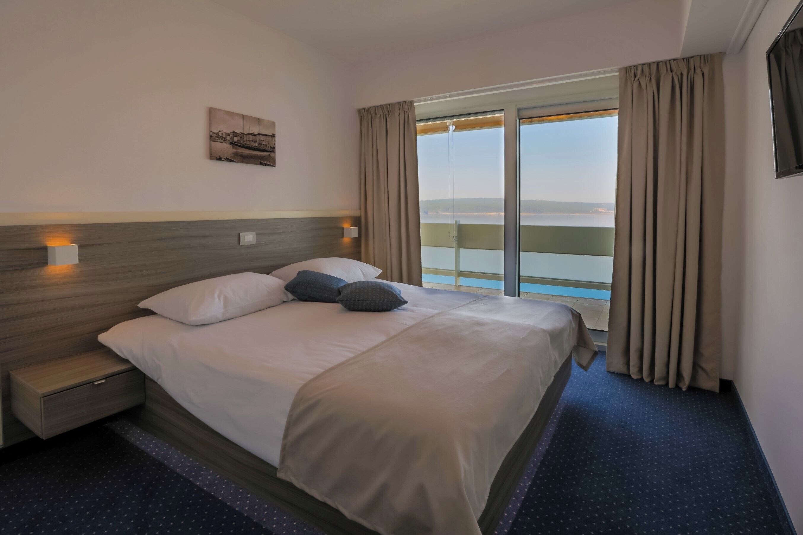 A hotel room with a bed, a nightstand, a window with curtains, and a view of the sea.