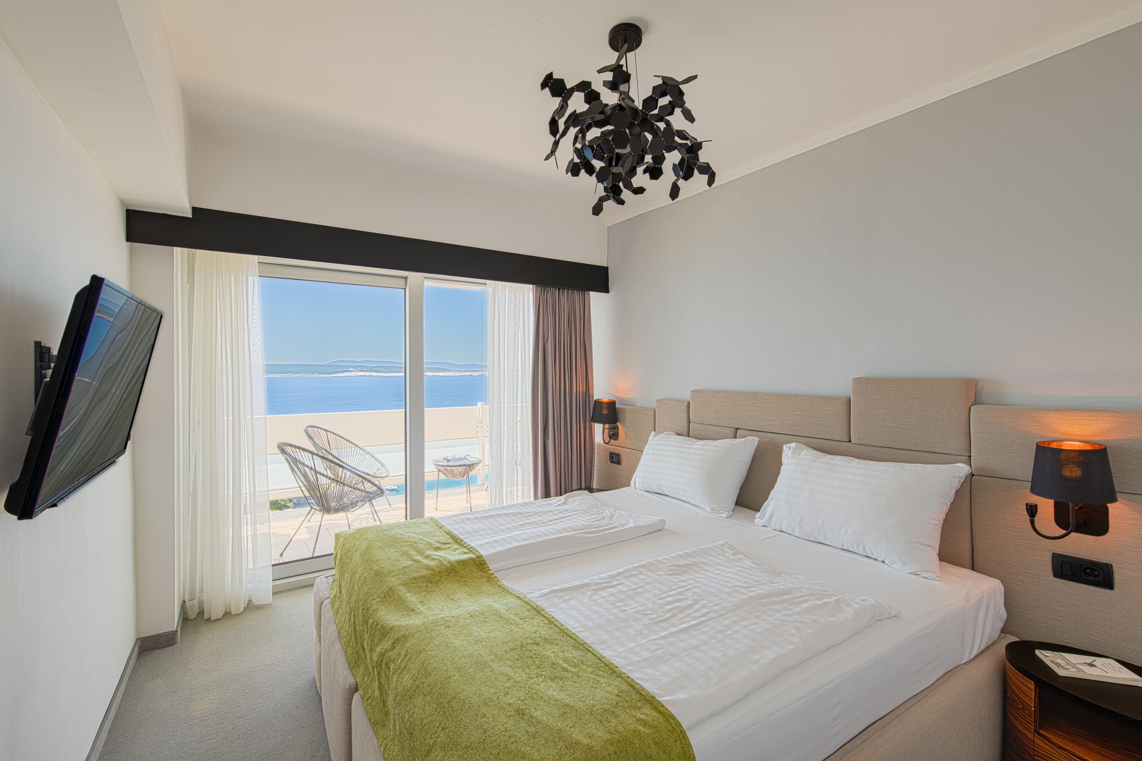 Une chambre d’hôtel avec un grand lit, un téléviseur fixé au mur, une fenêtre donnant sur la plage et un luminaire au plafond.