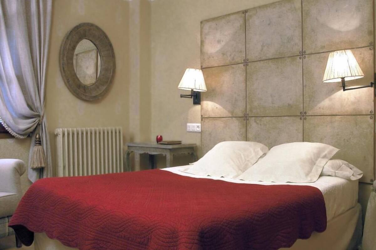 A hotel room with a bed, two bedside lamps, a radiator, a round mirror, and a small table with a book and an apple.