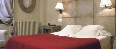 A hotel room with a bed, two bedside lamps, a radiator, a round mirror, and a small table with a book and an apple.