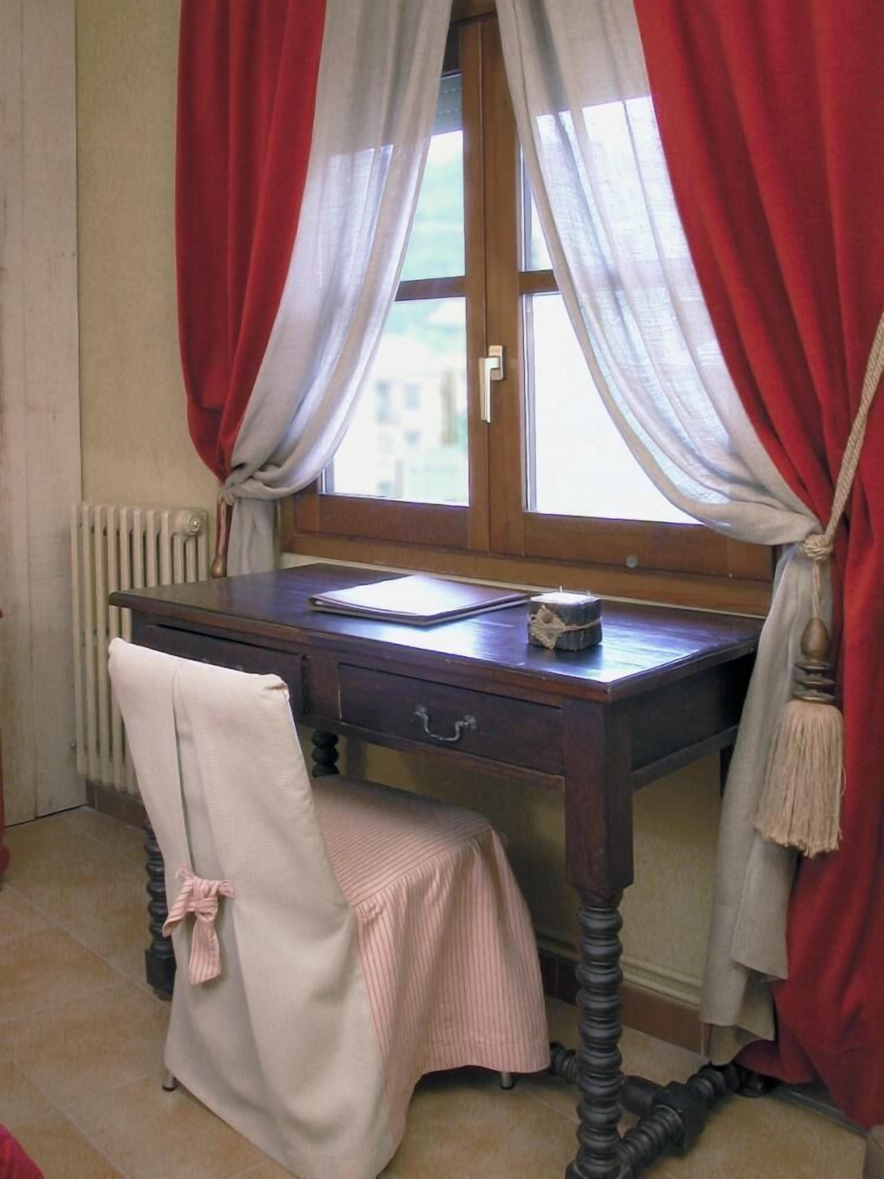 Una habitación con un escritorio de madera, una silla, un ventanal con cortinas rojas y blancas y un radiador.