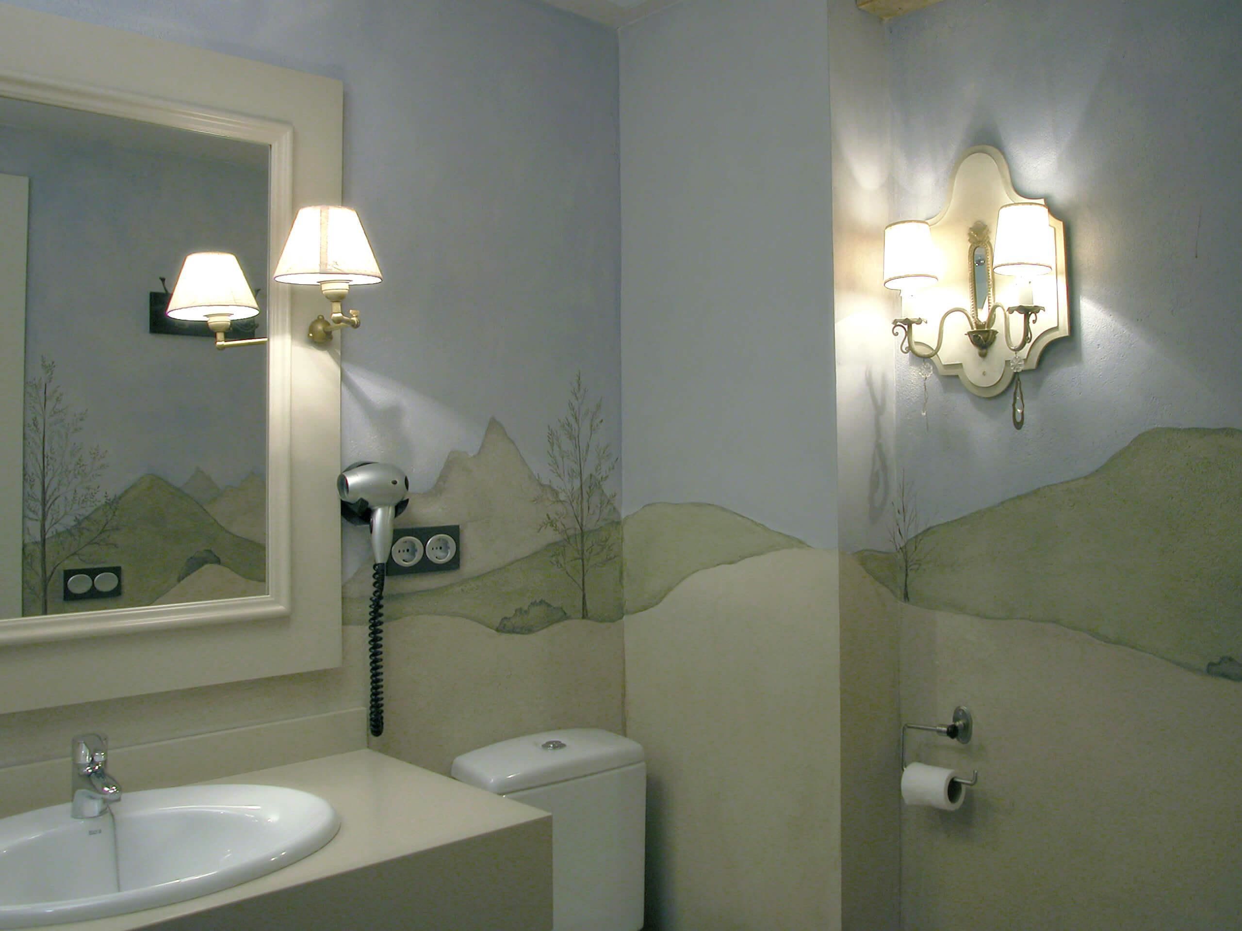 Un baño con lavamanos, inodoro y espejo con un mural de paisaje. Hay luces empotradas en la pared y un secador de pelo.