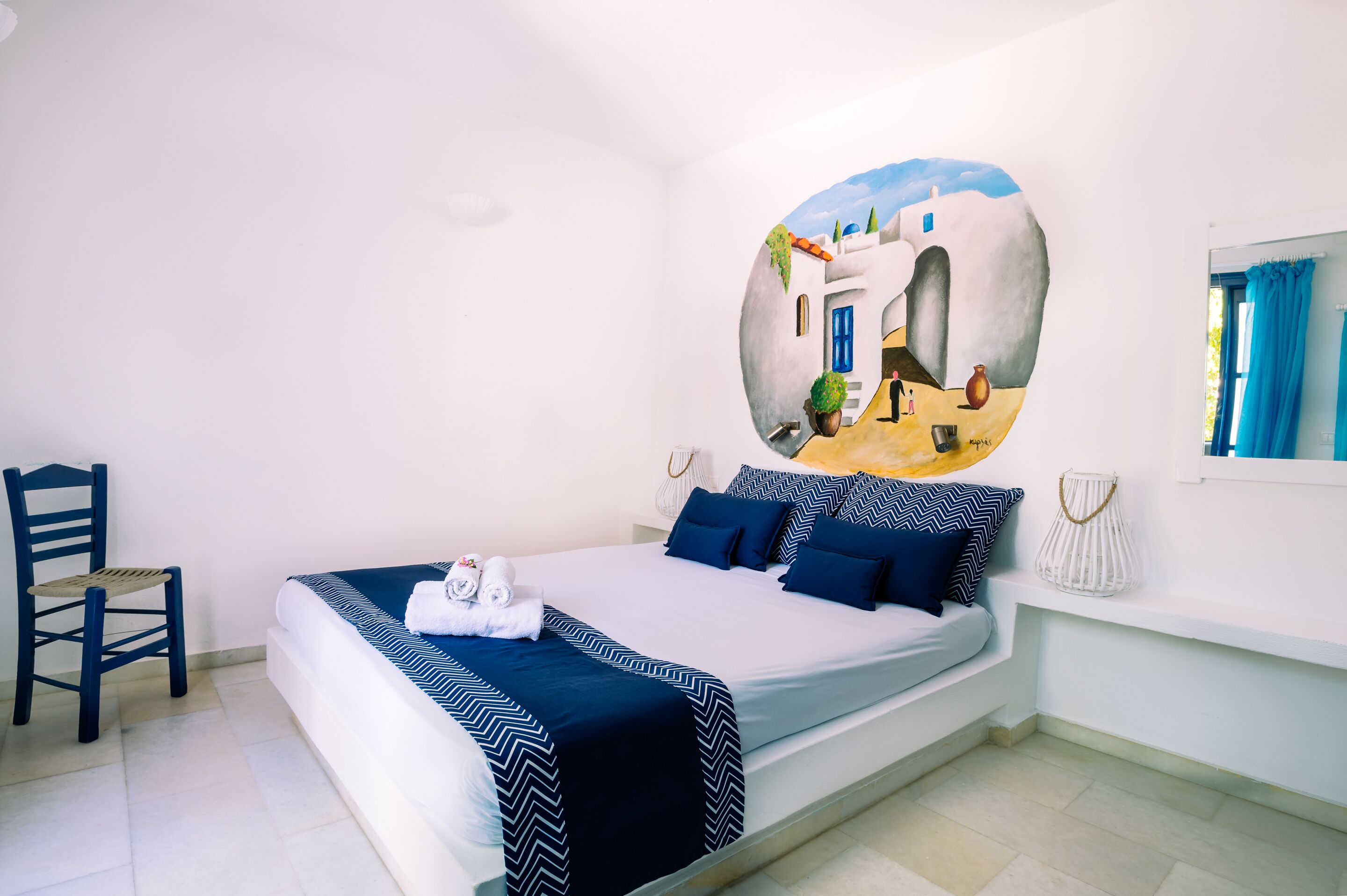 Una cama con una colcha de diseño azul y blanco, una silla, un mural en la pared y una ventana con cortinas.