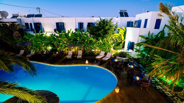 Una piscina amb gandules i taules, envoltada de plantes tropicals i edificis blancs amb accents blaus.