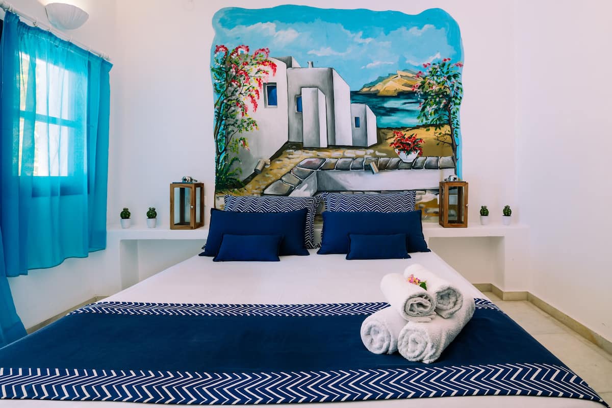 A hotel room with a bed, blue and white bedding, a painting of a coastal scene, and a window with blue curtains.