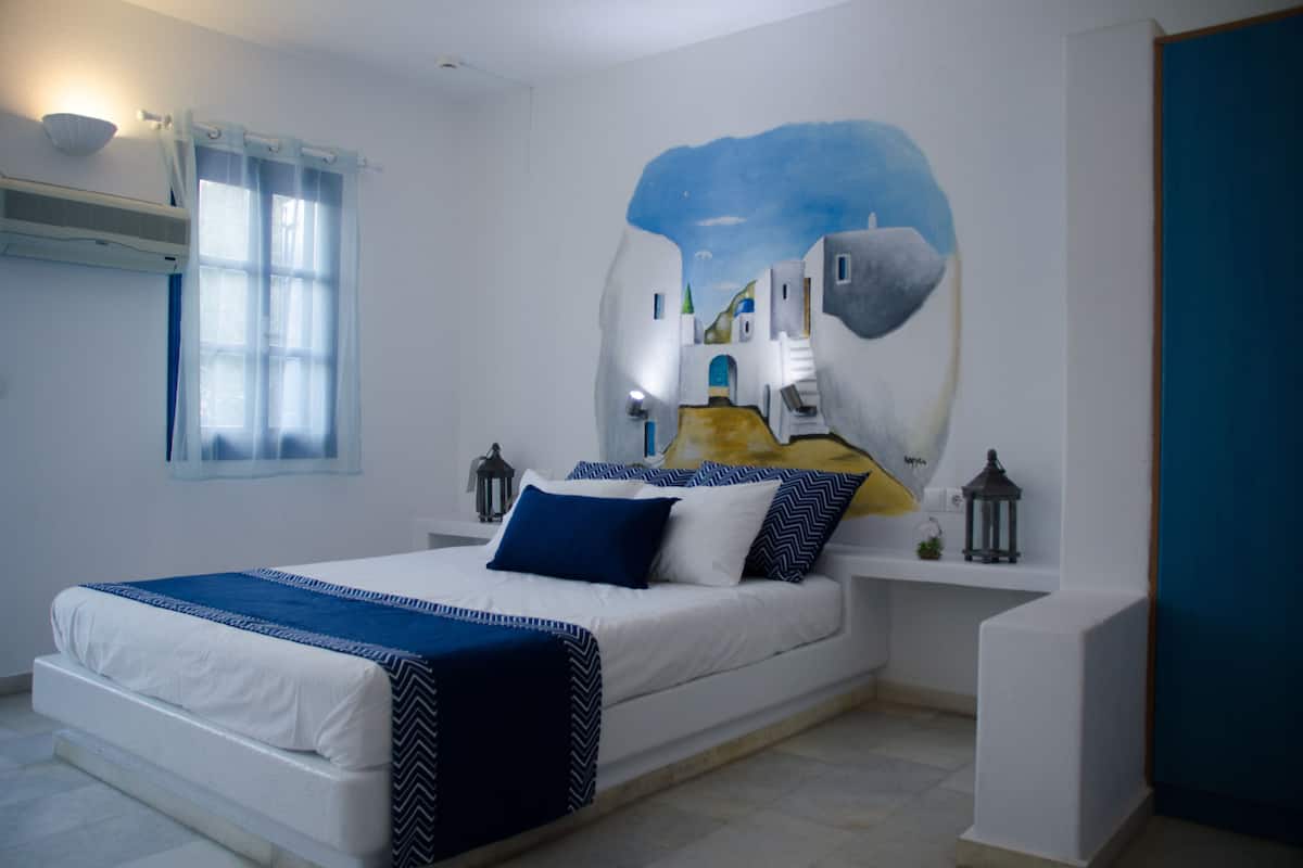 A bedroom with a bed, a nightstand, a wall-mounted air conditioner, a window with curtains, and a wall painting.
