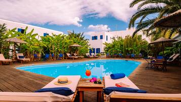Un área al lado de la piscina con sillones de descanso, una terraza de madera y una piscina azul cristalina.