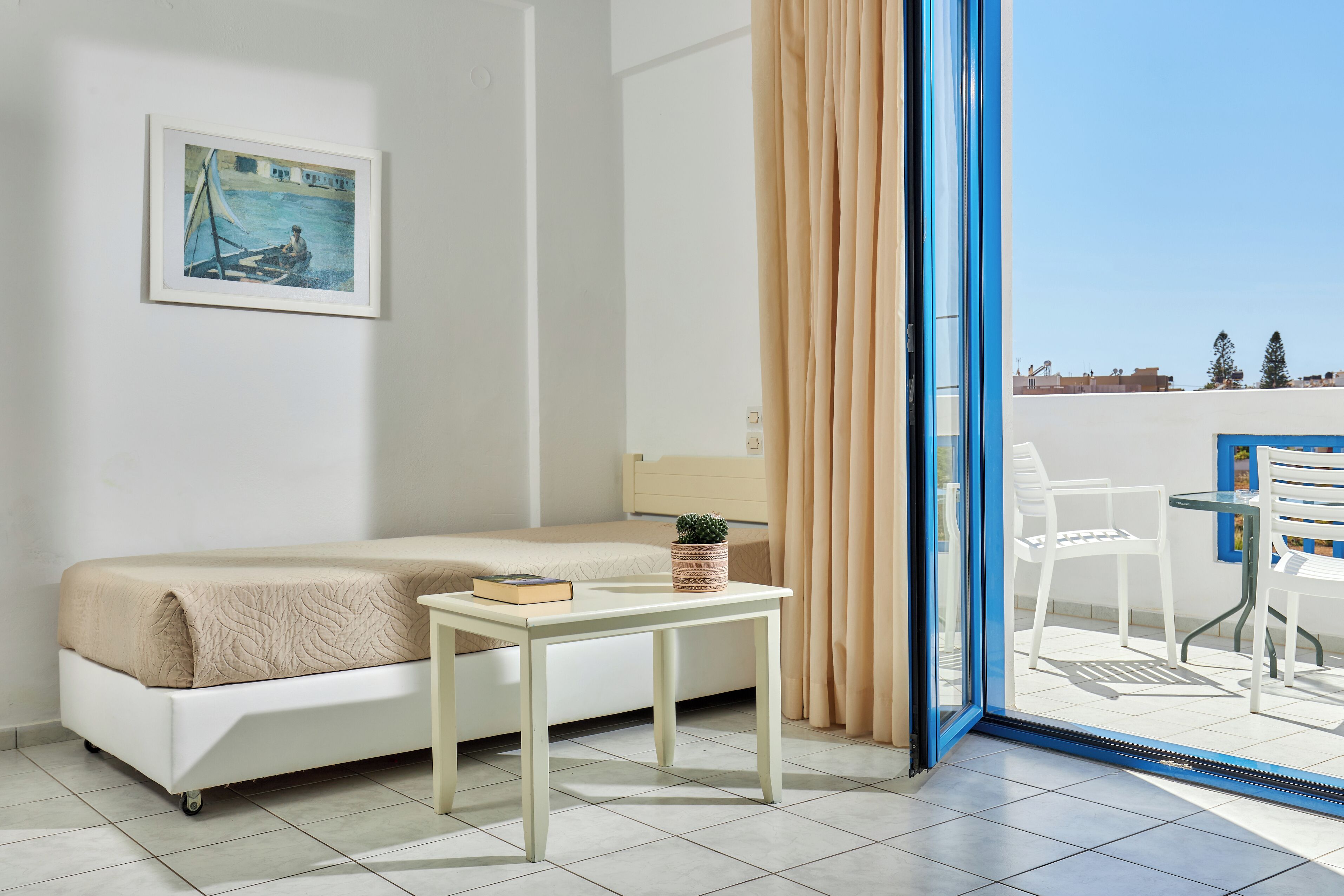 A room with a bed, a small table with a book and a plant, a painting on the wall, and a view of a balcony with chairs and a table.