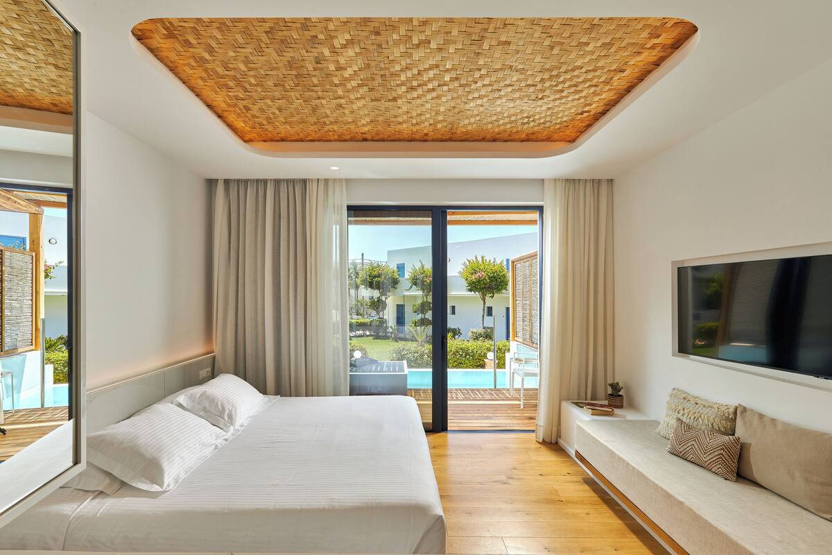 A bedroom with a bed, a sofa, a TV, and a view of a pool and greenery.