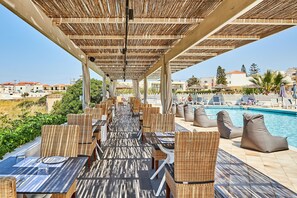 A covered outdoor dining area with wicker furniture and a pool in the background. - Hara Ilios Village (Hersonissos)