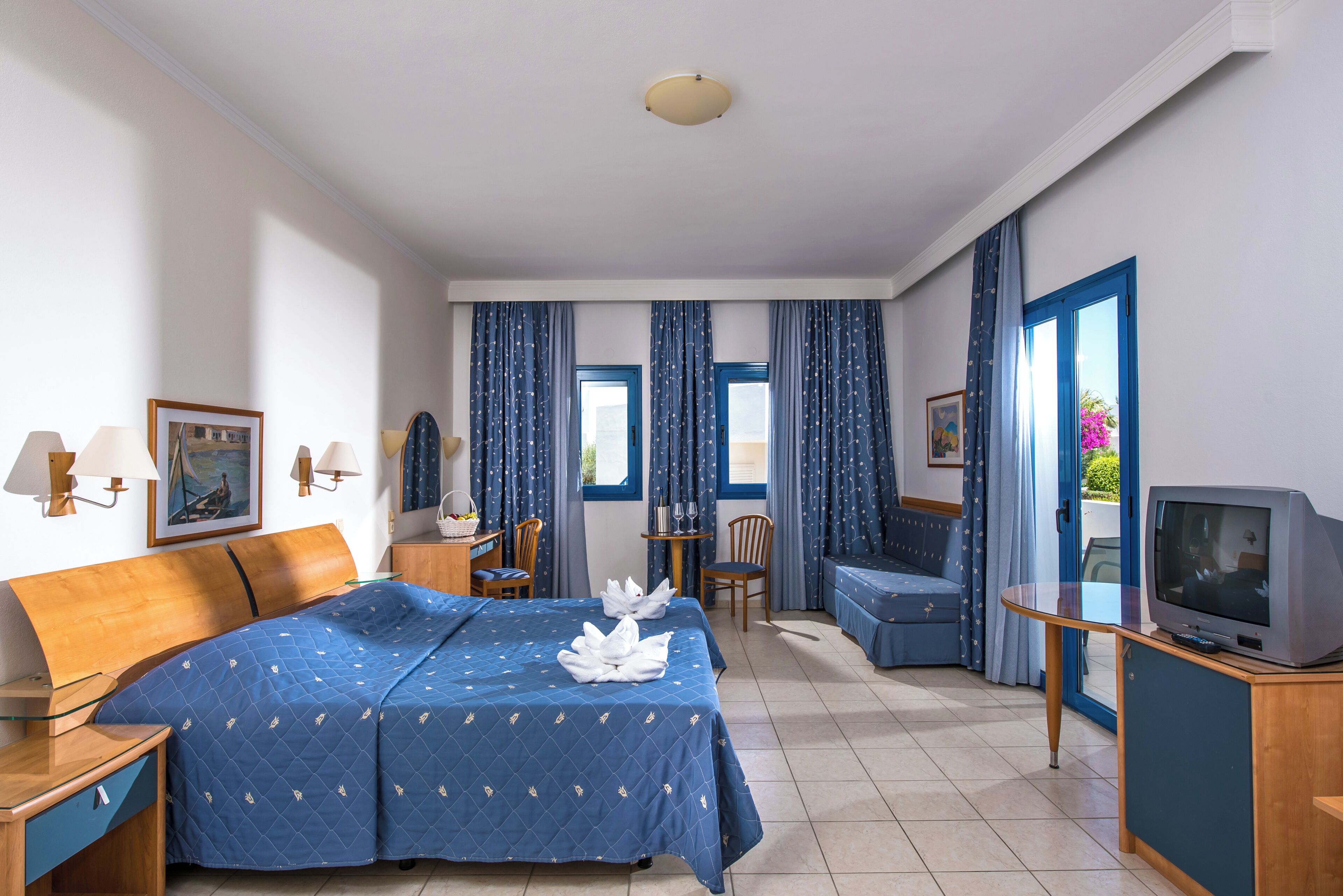 A hotel room with a large bed, a TV, a desk with a lamp, a chair, a small table, and a window with curtains.