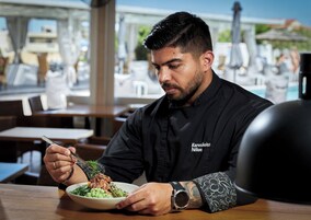 Una persona sentada a una mesa con un plato de comida, vistiendo un mandil de chef negro con texto.