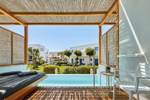 A balcony with a wooden floor, a white chair, a small table with a plant, and a view of a pool and buildings in the background. - Hara Ilios Village (Hersonissos)