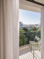 Varanda com vista para a paisagem urbana, guarda-corpo de metal e uma cadeira de metal verde.