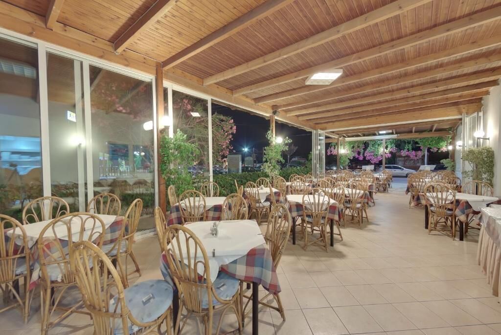 An outdoor dining area with wooden tables and chairs, covered by a wooden roof, and large windows on one side.