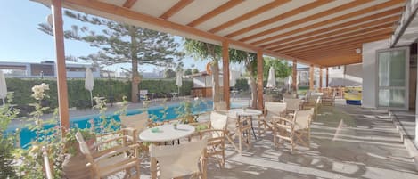 An outdoor patio area with wooden tables and chairs, a pool, and a covered seating area.