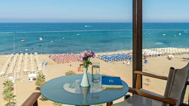 A table with a vase of flowers and a glass of water, overlooking a beach with umbrellas and lounge chairs. - Kriti Beach (Rethymno)