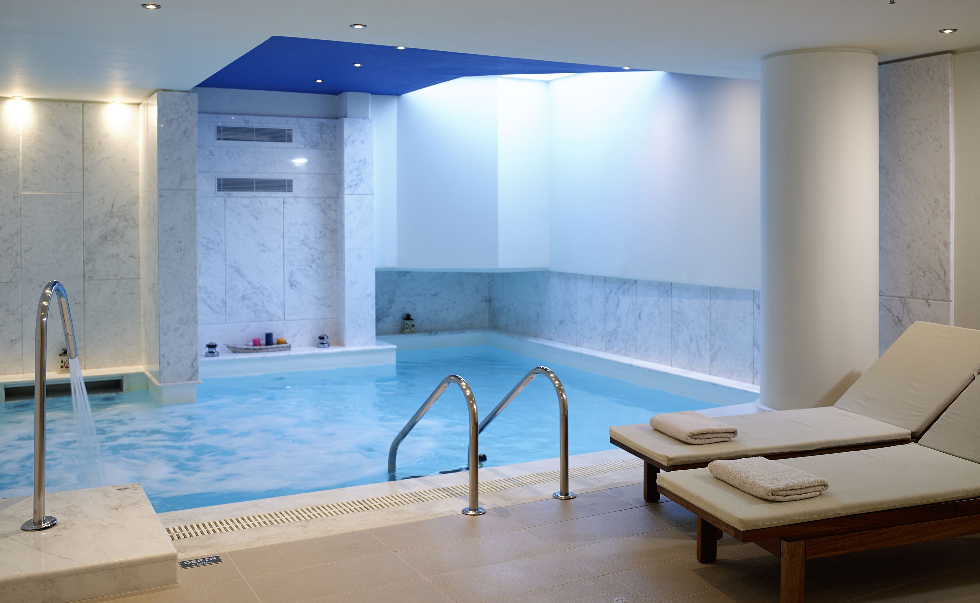 A modern indoor pool with a blue and white color scheme, a jacuzzi, and two lounge chairs with towels.