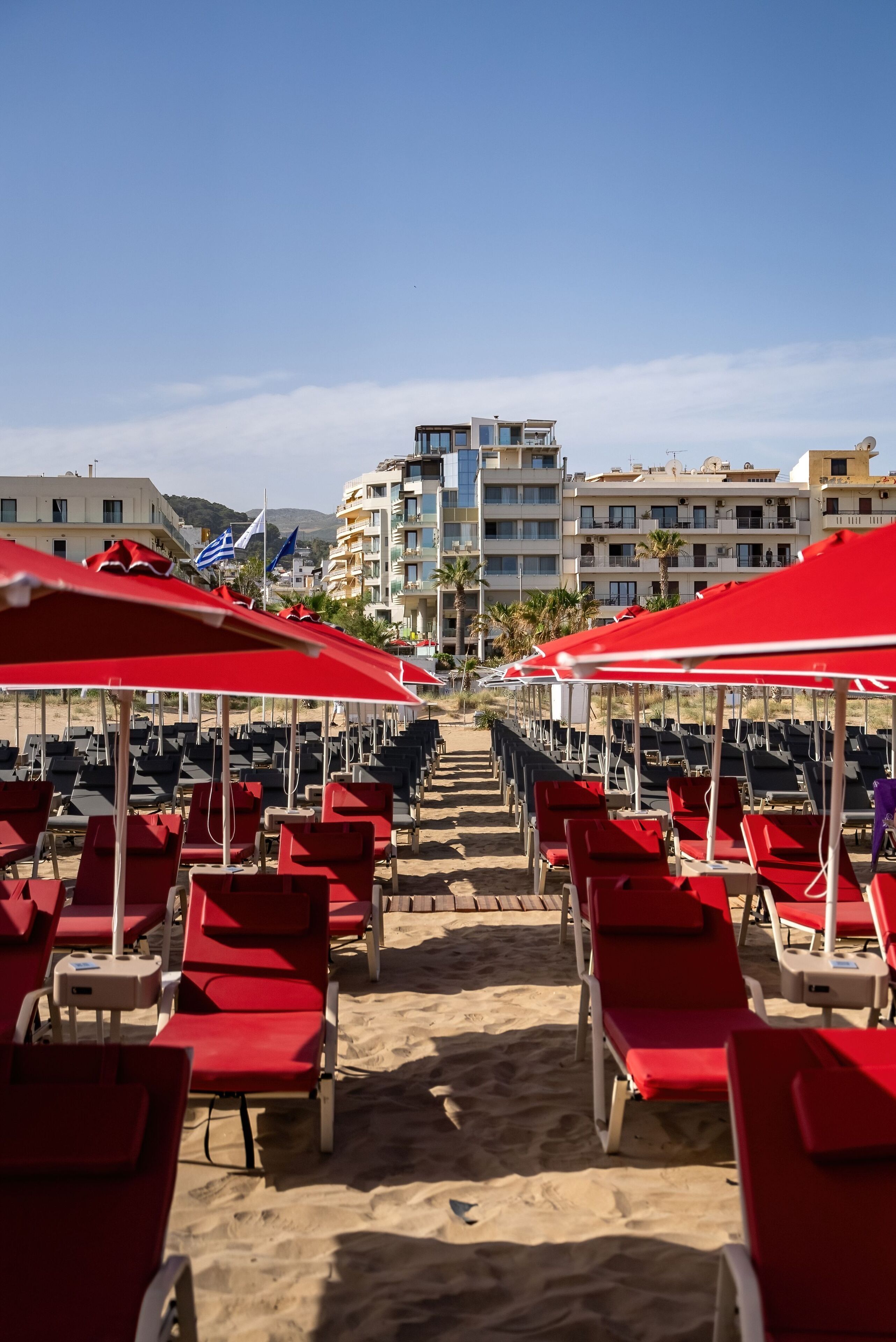 A beachside hotel with rows of red lounge chairs under red umbrellas, a clear sky, and buildings in the background.