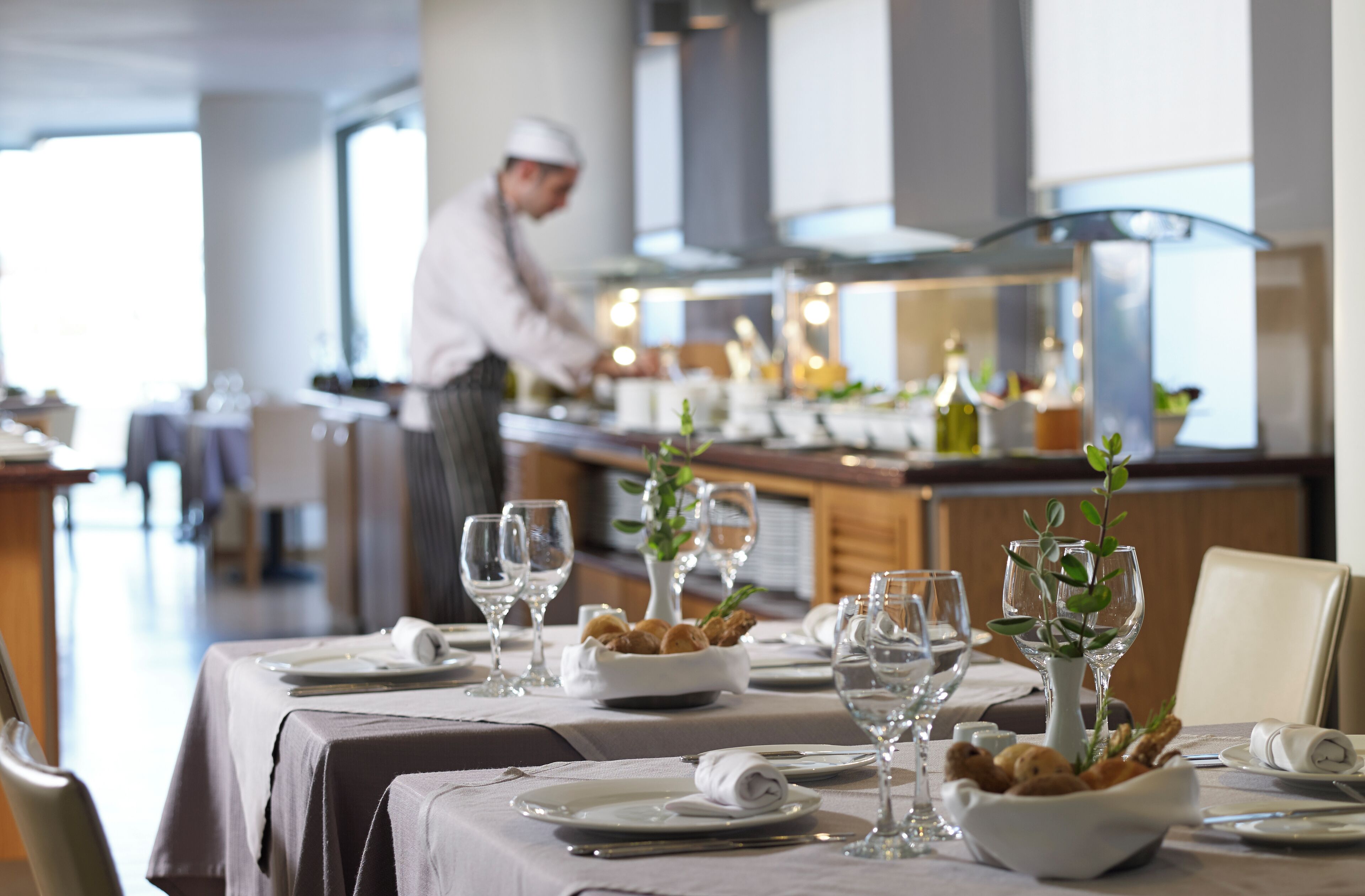 Comedor de restaurante con mesas preparadas para una comida, un chef al fondo y una cocina moderna a la vista.
