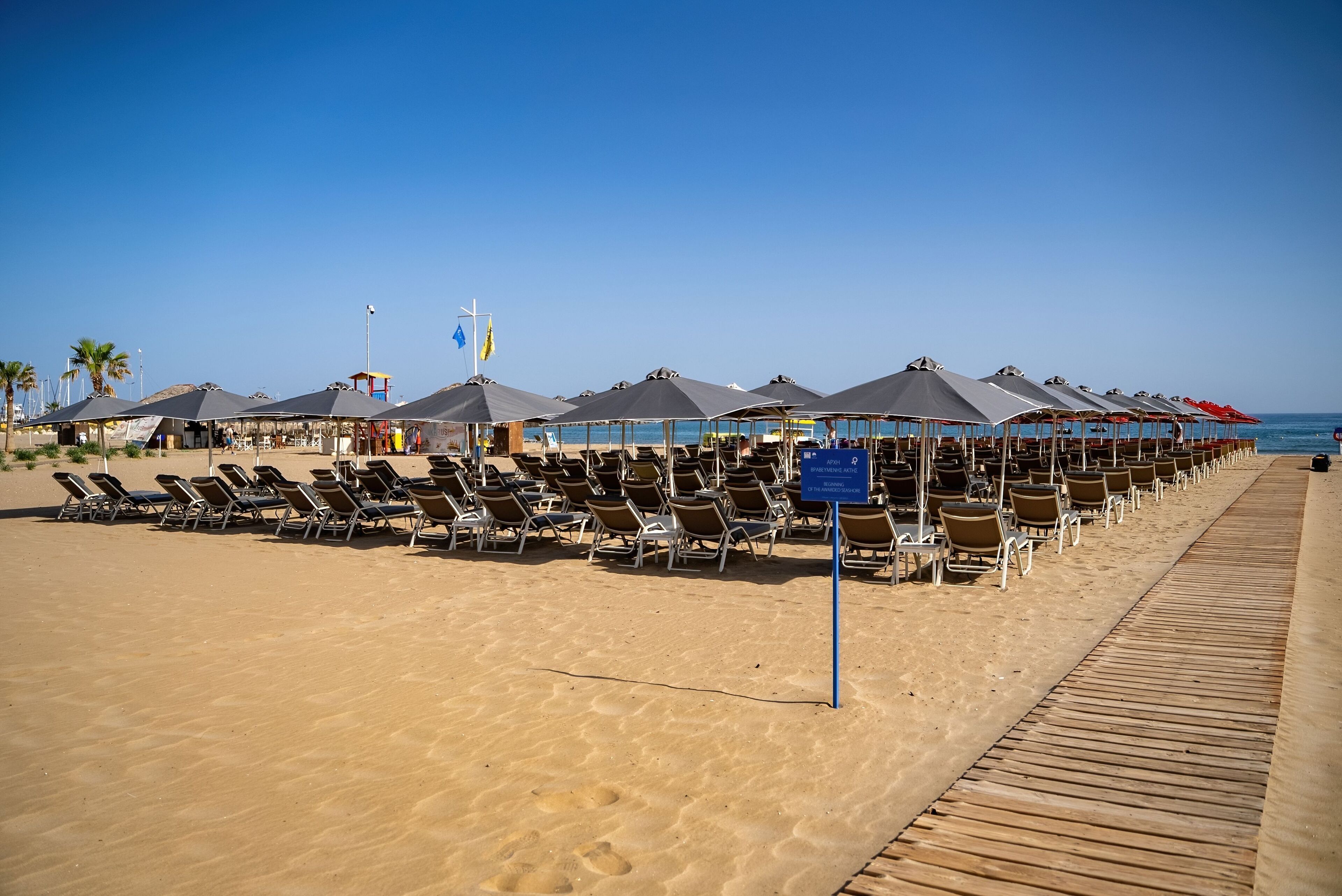 Platja amb files de gandules i ombrel·les, una passera de fusta i un senyal que indica que la platja forma part d'una costa guardonada.