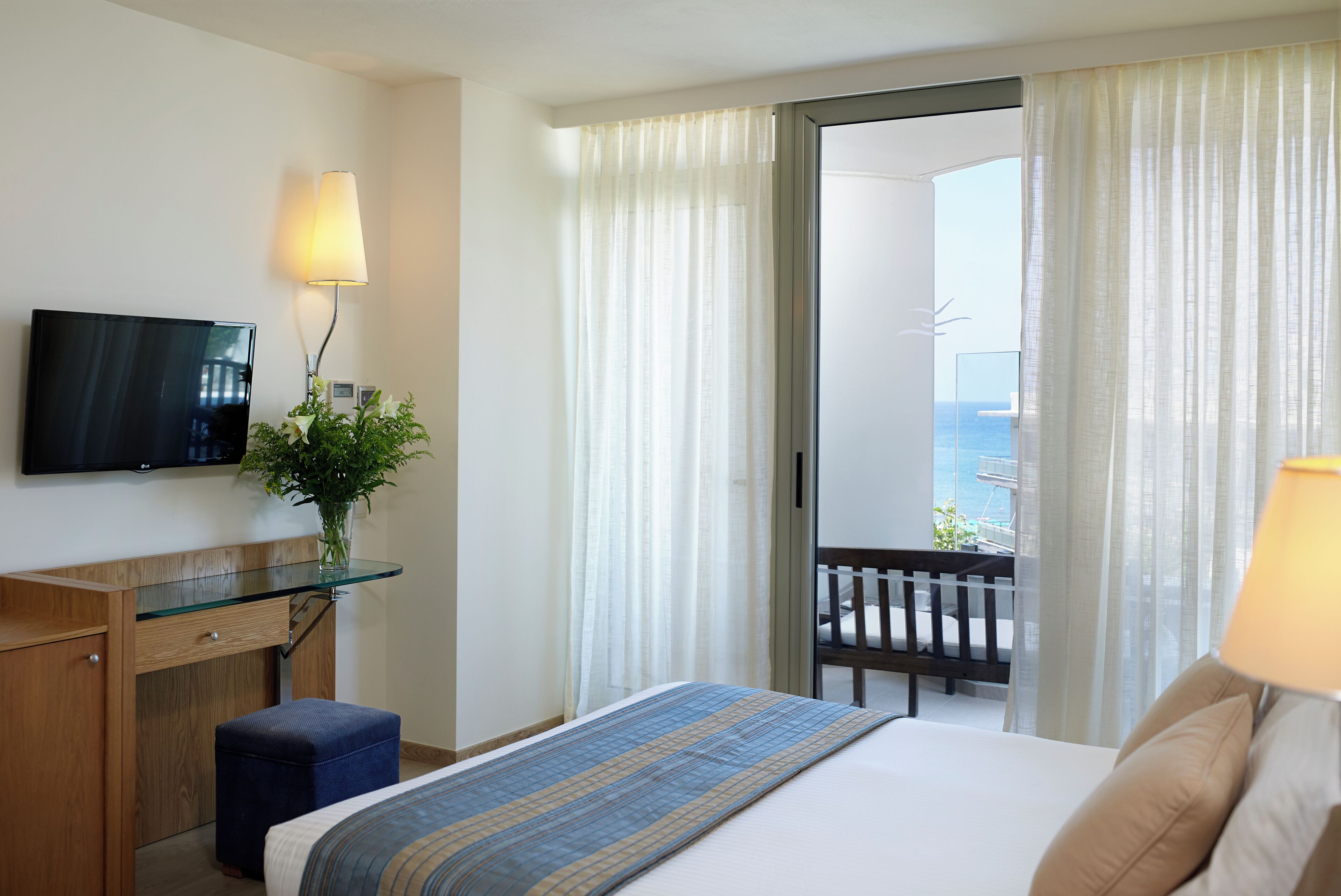 A hotel room with a bed, a desk with a TV, a window with curtains, and a view of the sea.