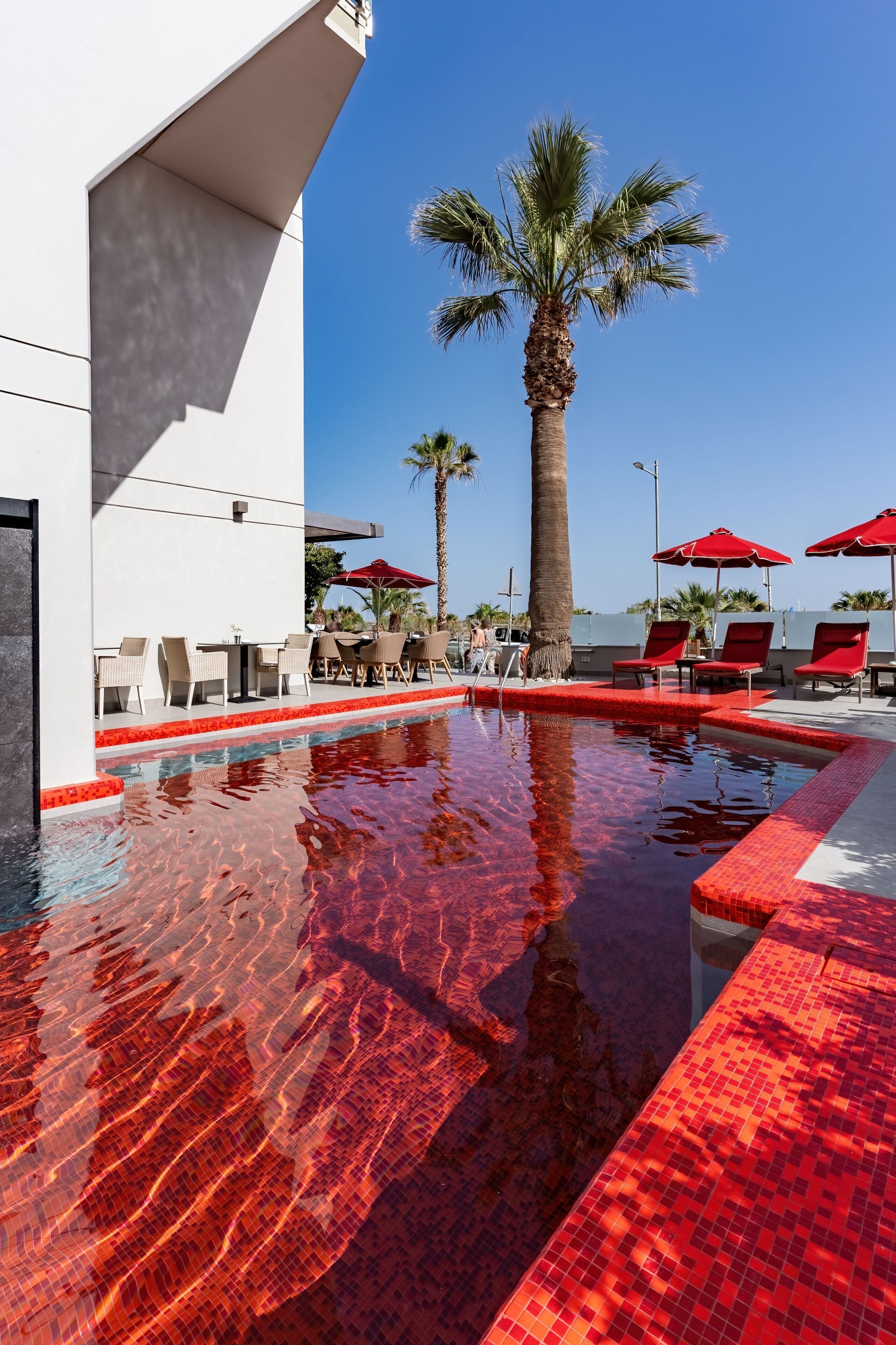 A swimming pool with red-tiled edges, surrounded by lounge chairs and umbrellas, and palm trees in the background.