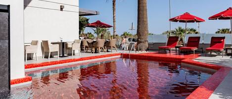 A swimming pool with red-tiled edges, surrounded by lounge chairs and umbrellas, and palm trees in the background.