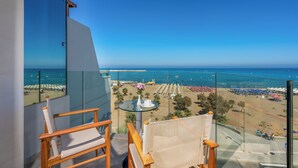 A balcony with two wooden chairs and a small table, overlooking a beach with umbrellas and people. - Kriti Beach (Rethymno)