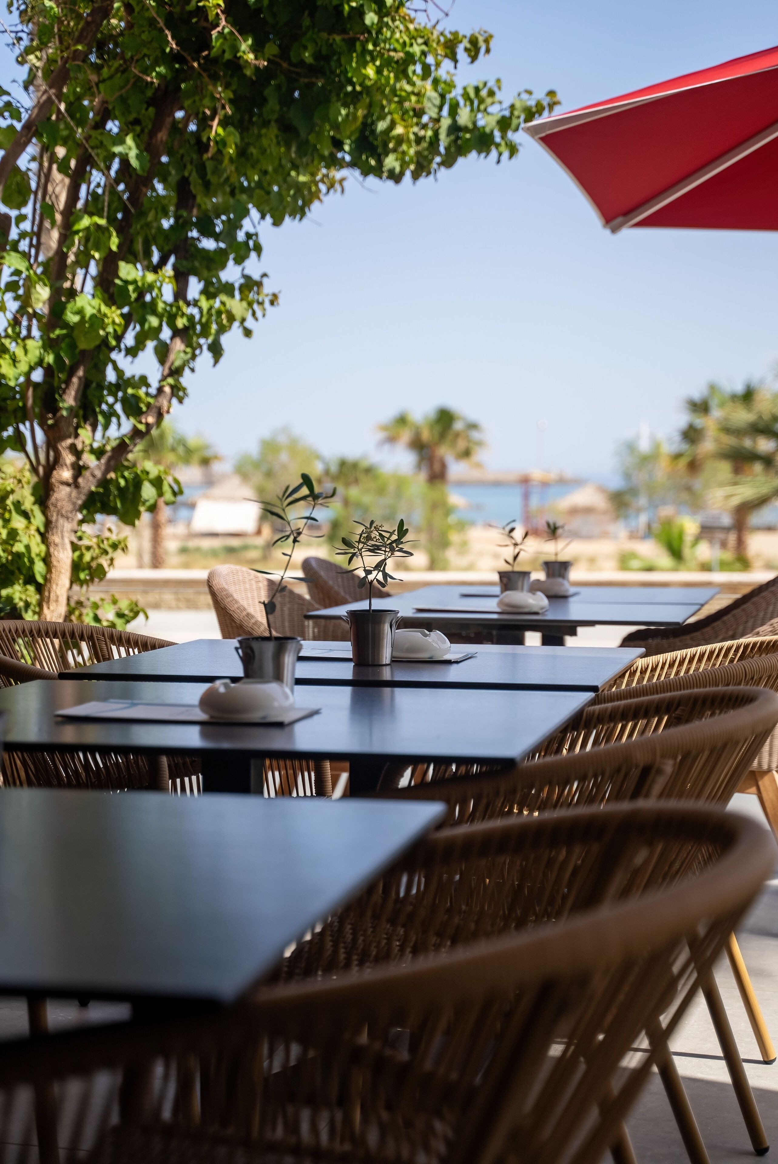 An outdoor dining area with wicker chairs and tables, a red umbrella, and a view of the beach and ocean.
