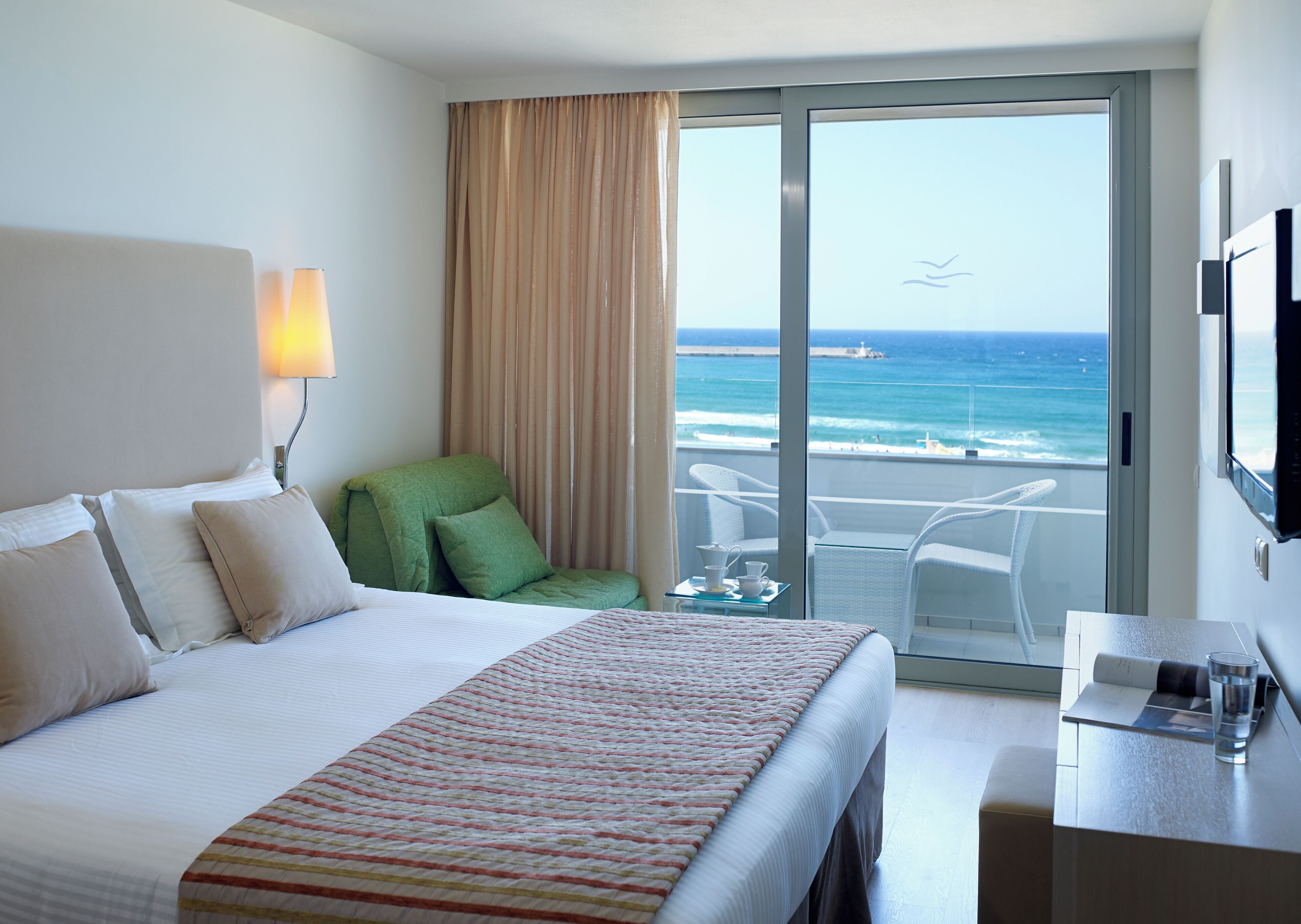Una habitación de hotel con una cama, una silla verde, un balcón con vista al océano y un televisor en la pared.