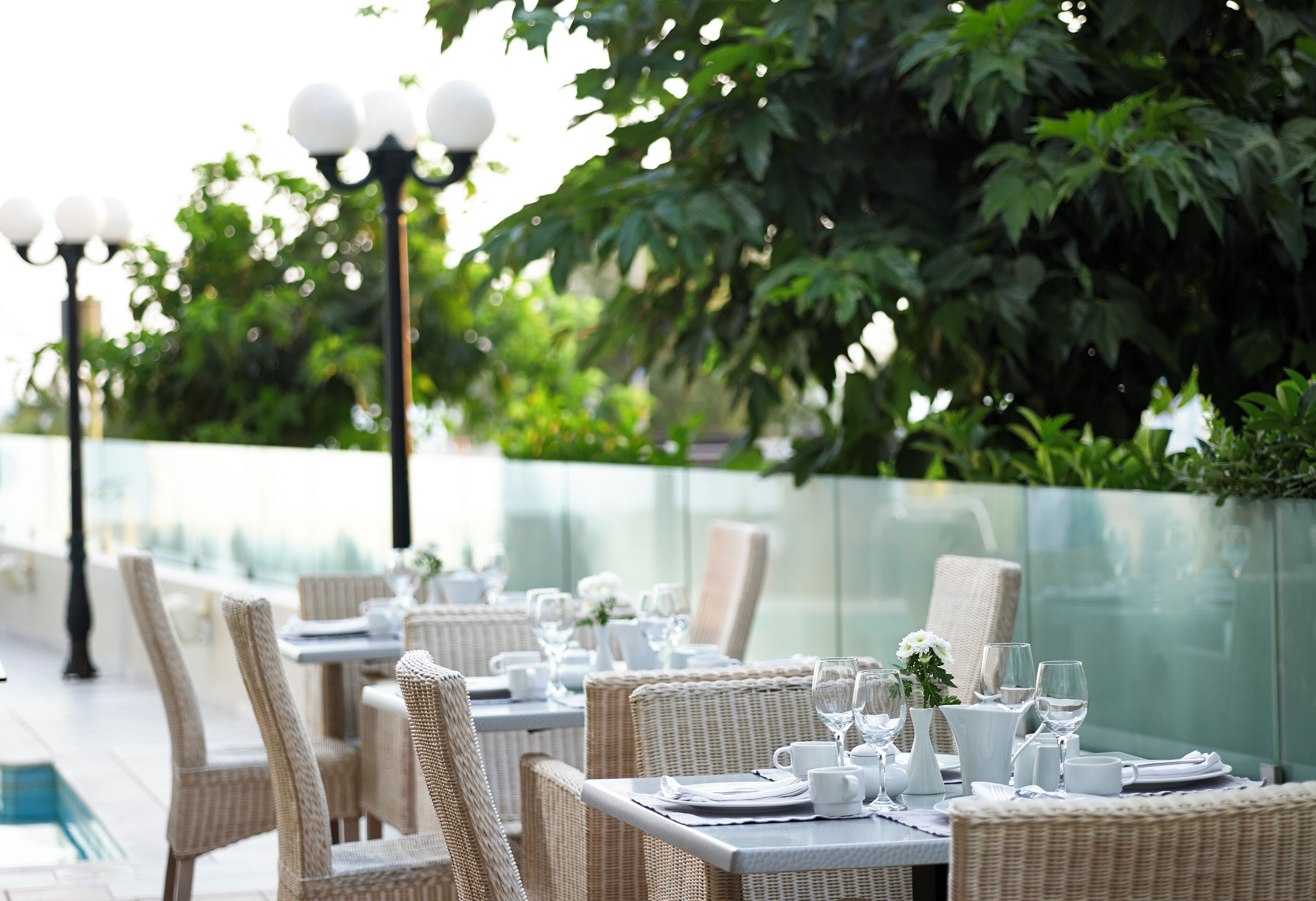 A dining area with wicker chairs and tables set for a meal, surrounded by greenery and a glass railing.