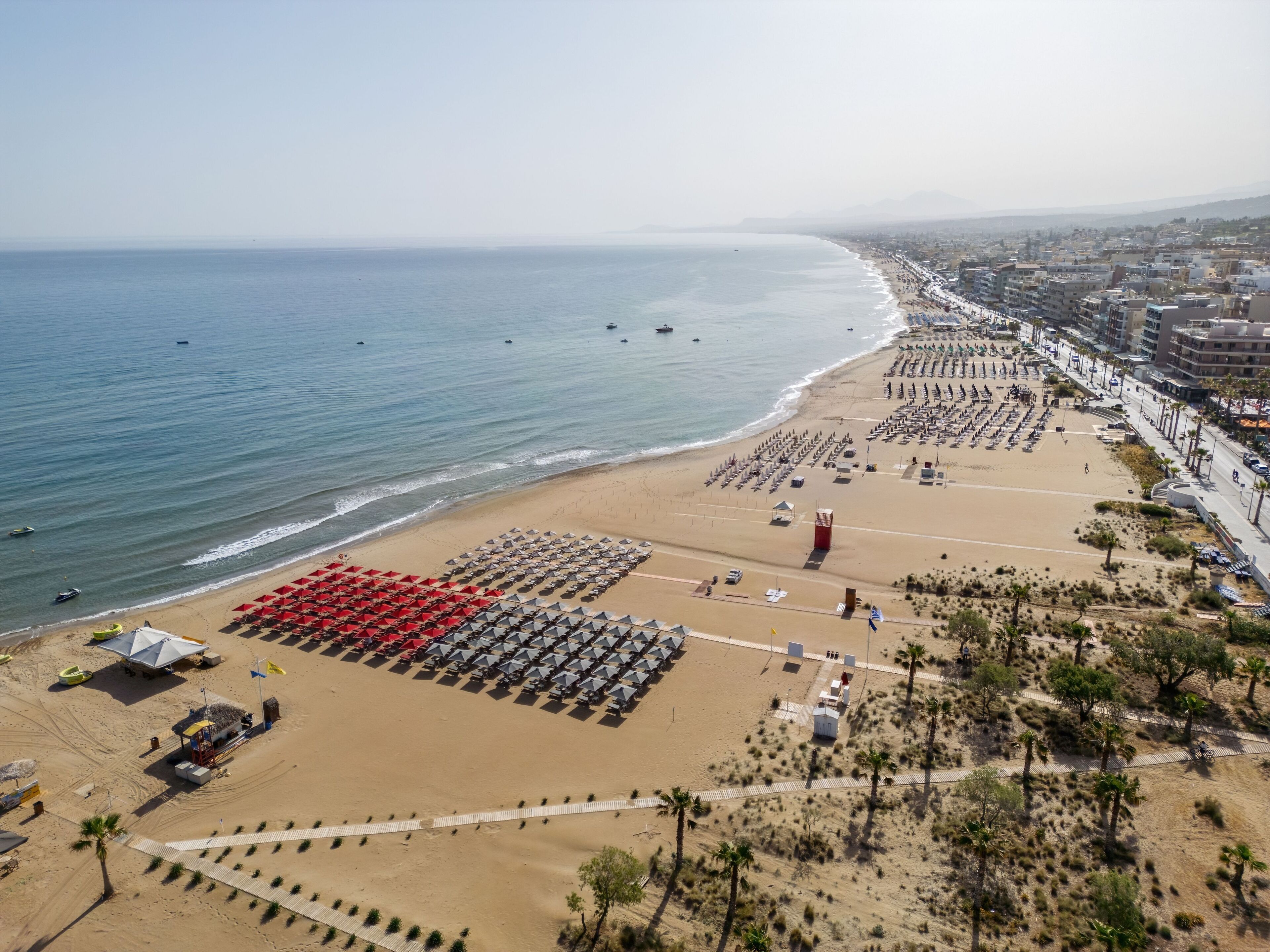 Una playa con hileras de sombrillas y tumbonas, una torre de salvavidas y un paseo marítimo que lleva a la playa.