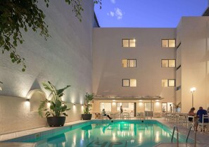 An indoor swimming pool with a tiled edge, surrounded by a building with multiple windows and a covered seating area.
