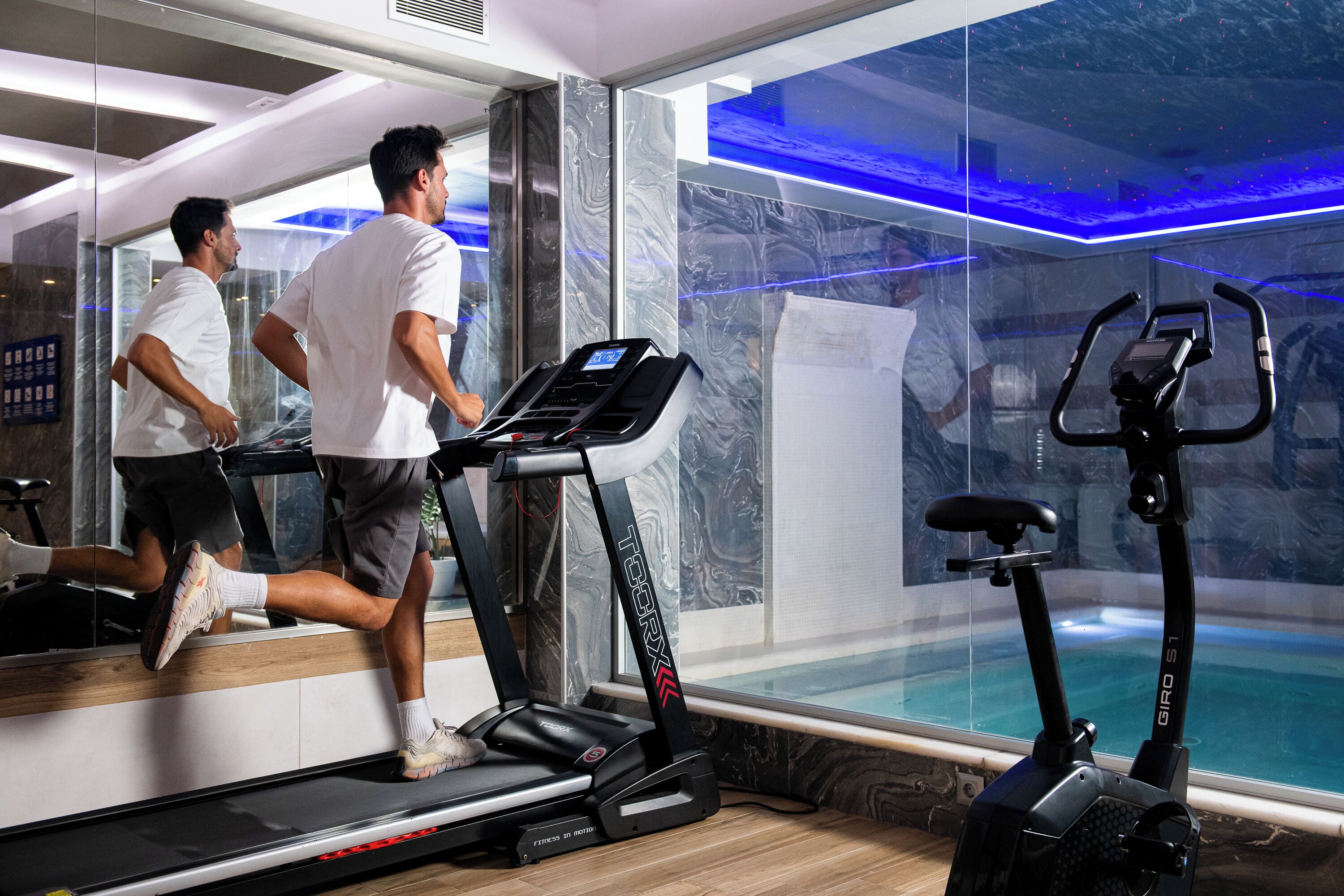A gym with treadmills and an elliptical machine, a large mirror, and a view of a swimming pool.
