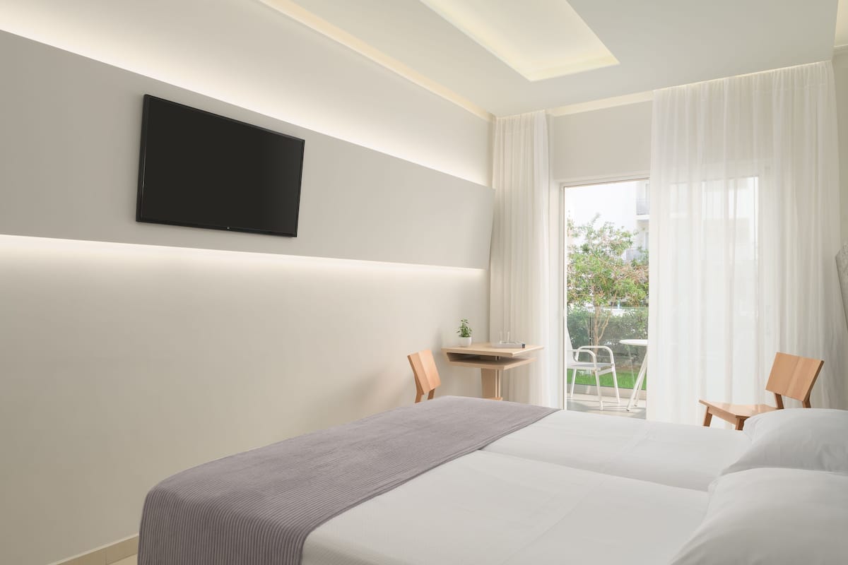 A hotel room with a bed, a TV mounted on the wall, a small table with a chair, and a view of the outdoors.