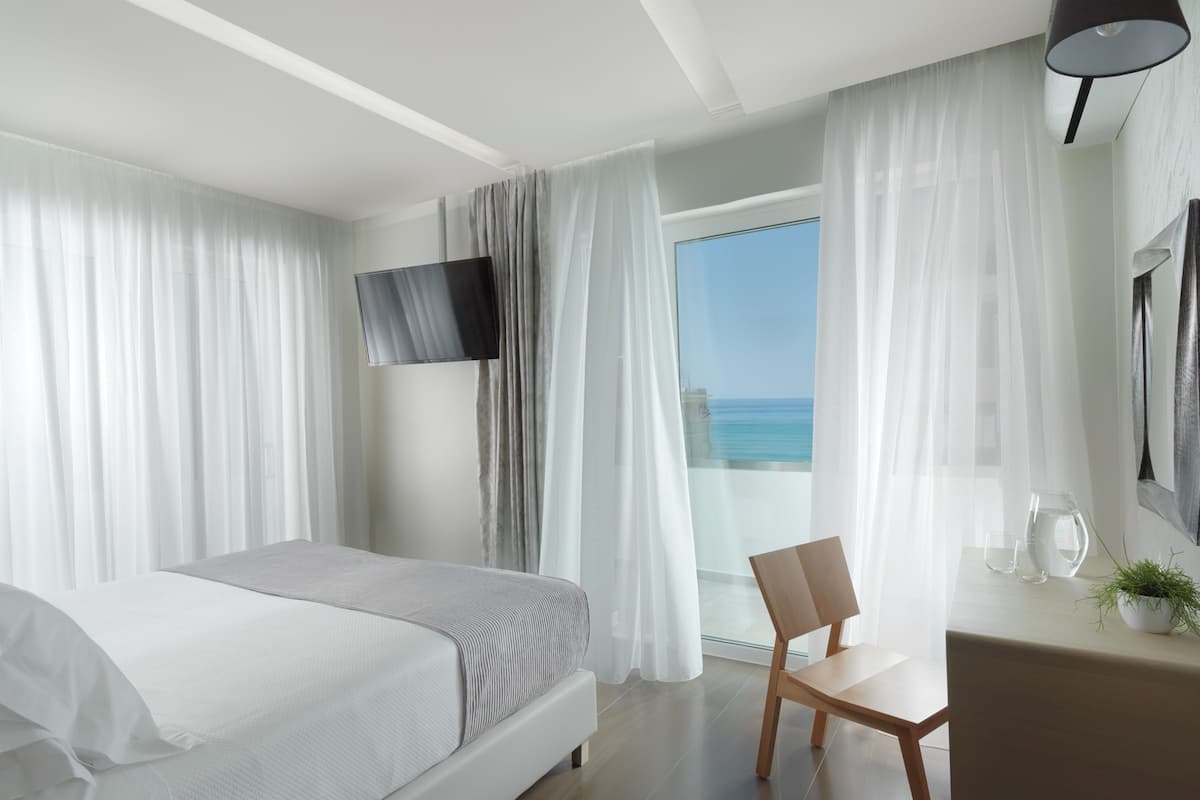 A modern hotel room with a bed, a desk, a chair, a TV, and a view of the sea.