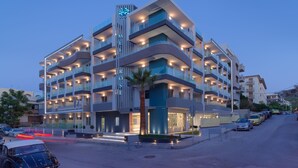 A multi-story hotel with balconies, a central entrance, and a parking area. - Melrose Rethymno by Mage Hotels (Rethymno)