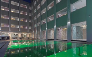 Interior moderno de un hotel con un área de piscina central, varios pisos con balcones y un frente de cristal que refleja la piscina.