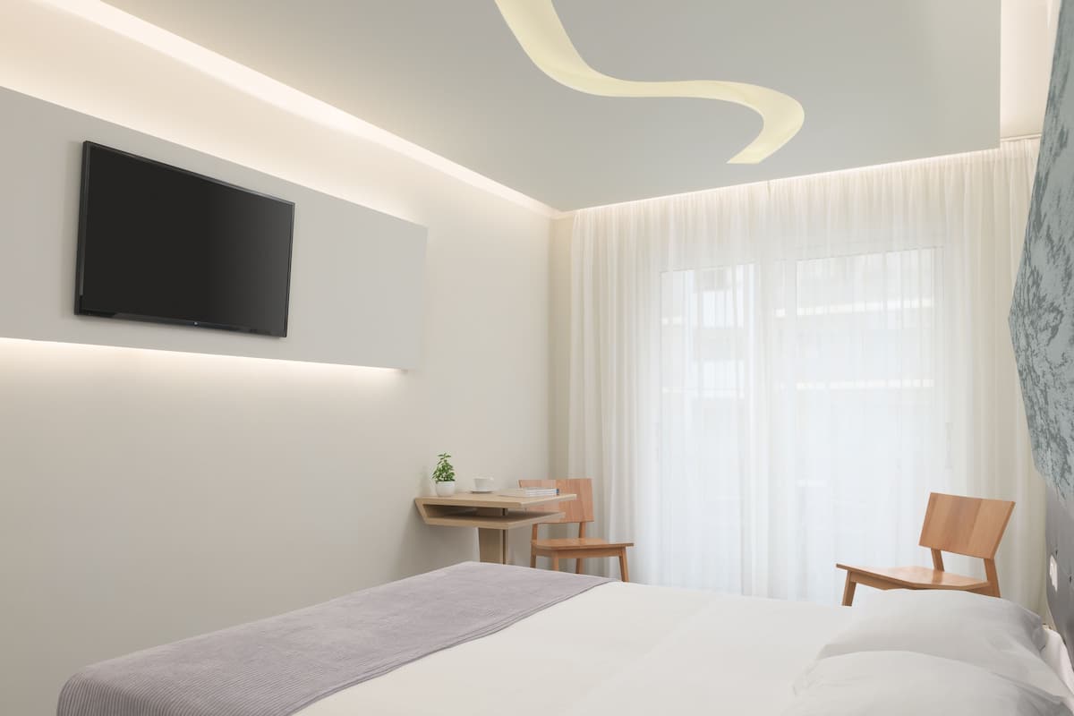 A modern hotel room with a bed, a desk, a chair, a television mounted on the wall, and a large window with sheer curtains.